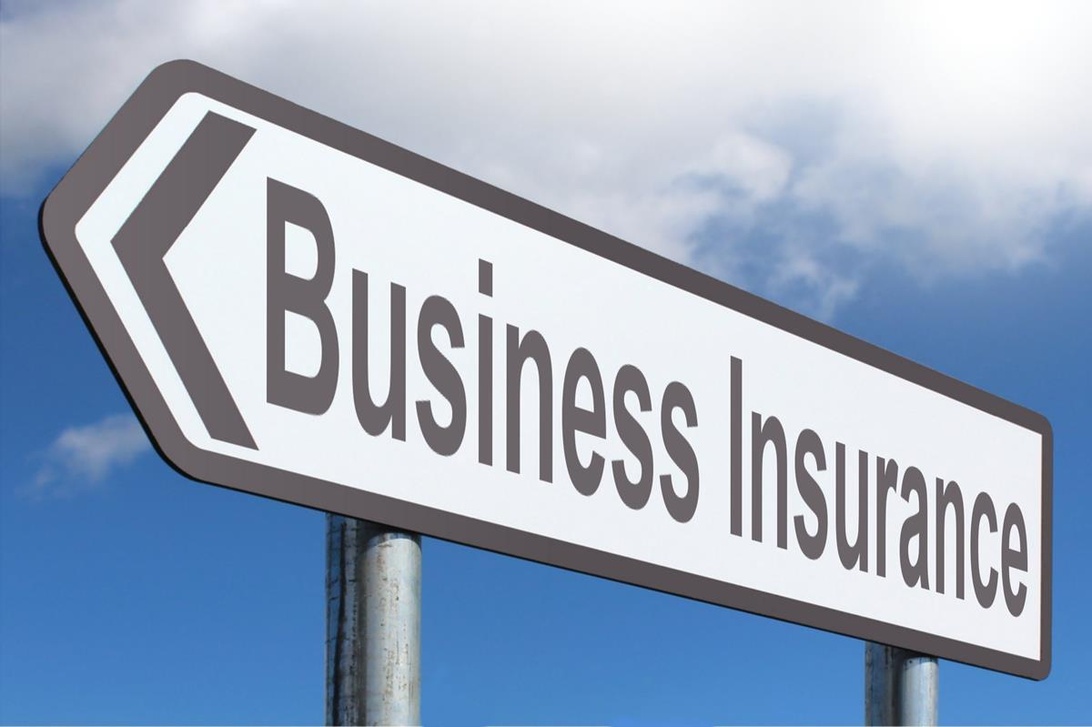 Et hvitt retningsskilt med teksten 'Business Insurance' mot en blå himmelbakgrunn. Skiltet er formet som en pil som peker mot venstre, og antyder at det gir retning til en tjeneste eller et kontor for bedriftsforsikring.
